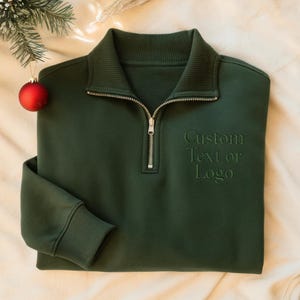 May include: A dark green sweatshirt with a quarter-zip closure and a ribbed collar. The text "Custom Text or Logo" is embroidered on the front. A red Christmas ornament hangs in the upper left corner.