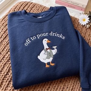 Könnte beinhalten: Ein marineblaues Sweatshirt mit Rundhalsausschnitt und einer Stickerei einer weißen Gans, die eine graue Weste und eine braune Fliege trägt. Die Gans hält einen Cocktailshaker und ein Martiniglas. Der Text "off to pour drinks" ist weiß gestickt.