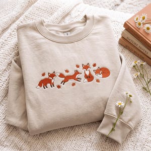 Könnte beinhalten: Ein beigefarbenes Sweatshirt mit Rundhalsausschnitt und einer Stickerei von vier Füchsen in verschiedenen Posen, darunter stehend, rennend, sitzend und schlafend, durchsetzt mit kleinen orangefarbenen Herbstblättern auf der Brust.