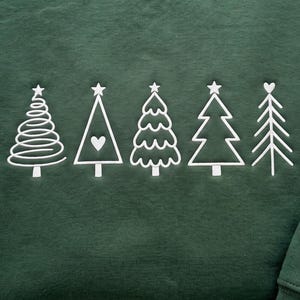 May include: A dark forest green sweatshirt featuring five white embroidered Christmas tree designs in a row. The trees include a spiral design, a triangle with a heart, a scalloped edge tree, a classic triangular tree, and a branch-style tree.
