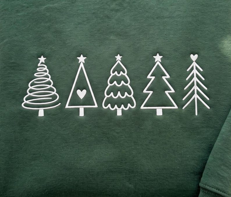 May include: A dark forest green sweatshirt featuring five white embroidered Christmas tree designs in a row. The trees include a spiral design, a triangle with a heart, a scalloped edge tree, a classic triangular tree, and a branch-style tree.