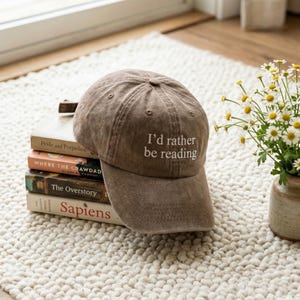 May include: A brown baseball cap with the text "I'd rather be reading" in white. The cap is resting on a stack of books, including "Pride and Prejudice" and "Sapiens". A small vase of flowers is in the background.