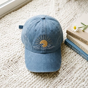 May include: A blue baseball cap with a sun and wave design embroidered on the front. The sun is yellow with gold rays, and the waves are white. The hat is sitting on a white, textured surface.