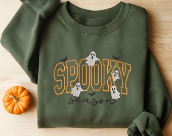 May include: An olive green sweatshirt with the words "SPOOKY season" in orange and white lettering. The design includes cartoon ghosts and bats, with a small orange pumpkin in the corner.