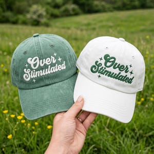 May include: Two baseball caps, one in muted green with white embroidery and one in white with dark green embroidery. Both caps feature the text "Over Stimulated" in a cursive font, accented by small four-pointed star icons on the front panels.