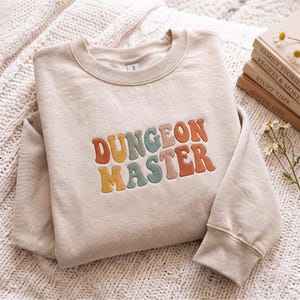 May include: A beige crewneck sweatshirt featuring the embroidered text "DUNGEON MASTER" in a wavy, multi-coloured font. The letters are coloured in shades of orange, yellow, teal, and muted pink, centred on the chest of the long-sleeved garment.
