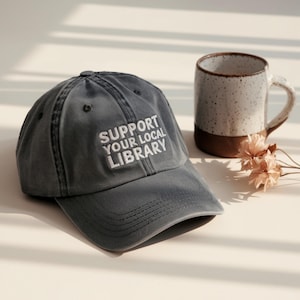May include: A gray baseball cap with the text "SUPPORT YOUR LOCAL LIBRARY" embroidered in white. A ceramic mug with a brown base and speckled design sits next to the cap. Dried flowers are also present.