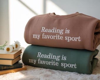 Könnte beinhalten: Zwei übereinander gestapelte Sweatshirts, eines braun und eines grün. Jedes hat den weißen, gestickten Text "Reading is my favorite sport". Die Sweatshirts sind gefaltet und wirken weich. Ein Stapel Bücher und Blumen im Hintergrund.