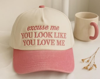 May include: A cream-colored baseball cap with a pink brim and pink embroidered text that reads, "excuse me YOU LOOK LIKE YOU LOVE ME." The cap features a structured crown and a button on top, showcasing a two-tone design with bold lettering.