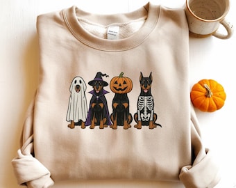 May include: Beige sweatshirt featuring four dogs dressed in Halloween costumes: a ghost, a witch, a jack-o'-lantern, and a skeleton. A small orange pumpkin and a mug are in the background. The sweatshirt is made of a soft material.