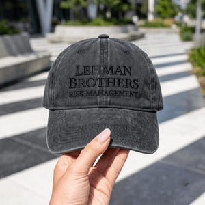 Può includere: Un cappellino da baseball grigio antracite con una finitura slavata e vissuta. La parte anteriore presenta la scritta ricamata in nero LEHMAN BROTHERS RISK MANAGEMENT in un carattere serif. Il cappello ha una visiera curva e sei pannelli.