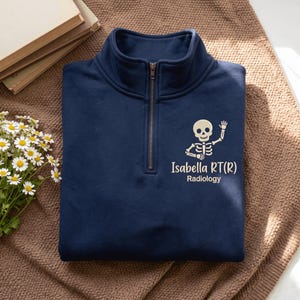 May include: A navy blue quarter-zip sweatshirt featuring a graphic of a white skeleton waving its hand. Below the skeleton, the text reads "Isabella RT(R)" and "Radiology" in a light-colored font. The garment has a metal zipper at the collar.