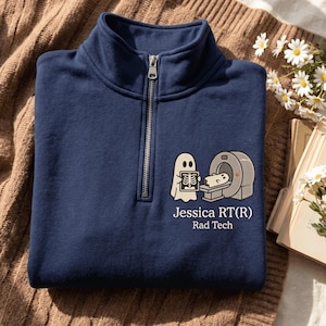May include: A navy blue quarter-zip sweatshirt featuring a graphic of a ghost operating an MRI machine on another ghost, with the text "Jessica RT(R) Rad Tech" printed below the illustration in a clean, white, sans-serif font.