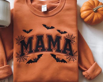 May include: An orange sweatshirt with the word "MAMA" in black, with black bats and spiderwebs. A small pumpkin is to the right of the sweatshirt. The sweatshirt is a fall-themed item.
