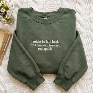 May include: An olive green crewneck sweatshirt featuring white embroidered text on the chest that reads, "i might be laid back but i can lean forward real quick." The garment has long sleeves, a ribbed collar, and ribbed cuffs.