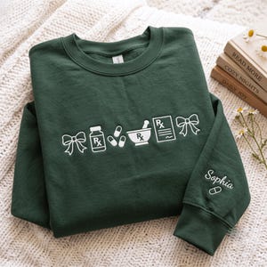 May include: A dark green crewneck sweatshirt featuring white embroidered icons of bows, a prescription bottle, pills, a mortar and pestle, and a prescription pad. The sleeve is embroidered with the name Sophia and a single pill icon.