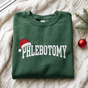 Puede incluir: Una sudadera de cuello redondo verde oscuro con texto bordado en blanco que dice PHLEBOTOMY en una fuente arqueada. Un pequeño gorro de Papá Noel rojo y blanco está bordado sobre la letra P, añadiendo un detalle festivo al diseño frontal.