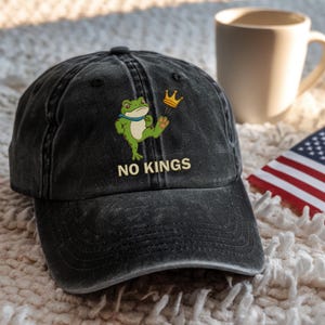 May include: Black baseball cap with an embroidered green frog holding a gold crown. The text "NO KINGS" is embroidered below the frog. The cap is sitting on a white surface.
