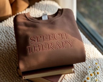 May include: A brown sweatshirt with the embroidered text "SPEECH THERAPY" in a lighter brown color. The sweatshirt is folded and resting on top of two books. The image is taken in natural light, with a cozy, inviting aesthetic.