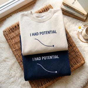 May include: Two crewneck sweatshirts, one cream-colored and one navy blue, are stacked on a woven basket. Each sweatshirt has the embroidered text "I HAD POTENTIAL" above a curved line design.