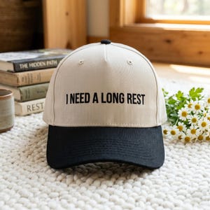 May include: A beige baseball cap with a black brim and the embroidered text "I NEED A LONG REST". The cap has a black button on top and is sitting on a white rug. Books and flowers are in the background.