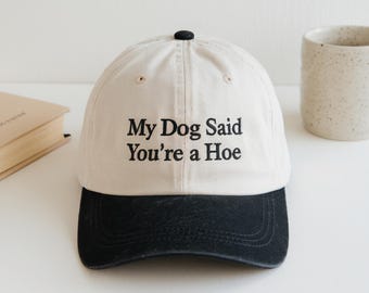 May include: A baseball cap with a cream-colored crown and a black brim. The front of the crown features black embroidered text that reads, My Dog Said You're a Hoe. The cap has a black button at the top center and stitched eyelets on the panels.