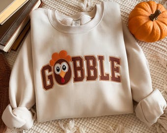 May include: Cream-colored sweatshirt with the word "GOBBLE" in brown varsity-style lettering. A cartoon turkey is embroidered on the front. An orange pumpkin sits to the right of the sweatshirt. The sweatshirt is folded on a cream-colored blanket.