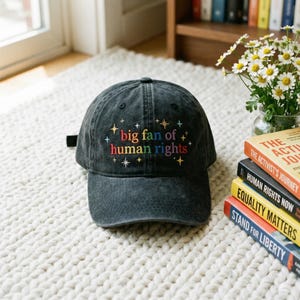 May include: A black baseball cap with the embroidered text "big fan of human rights" in colorful letters. The cap is sitting on a white, textured surface, with books and flowers in the background.