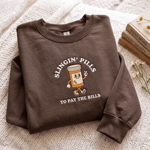 May include: A dark brown crewneck sweatshirt featuring a graphic of a cartoon pill bottle with a face, arms, and legs. The text reads "SLINGIN' PILLS TO PAY THE BILLS" in white lettering. The pill bottle has an orange body, white cap, and an Rx symbol.