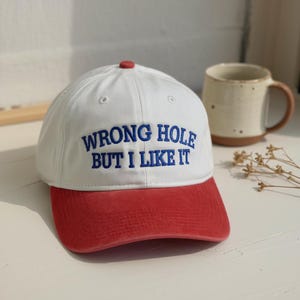 May include: A white baseball cap with a red brim and button. The text "WRONG HOLE BUT I LIKE IT" is embroidered in blue on the front. A casual, everyday accessory.