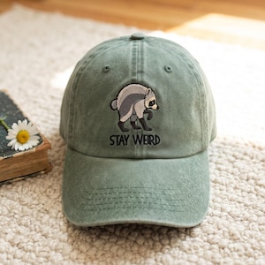 Puede incluir: Una gorra de béisbol color verde salvia con un bordado de un mapache gris y blanco. Debajo del mapache, el texto STAY WEIRD está bordado en hilo negro. La gorra tiene una corona estructurada, una visera curva y un botón en la parte superior.