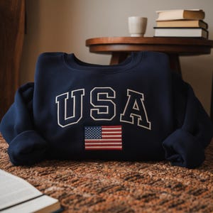May include: A navy blue sweatshirt with the word "USA" in white block letters and a small embroidered American flag. The sweatshirt is folded on a brown, textured surface.