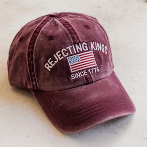 May include: A maroon baseball cap with the text "REJECTING KINGS" and an American flag embroidered on the front. The text "SINCE 1776" is below the flag. The cap has a curved brim and a distressed look.