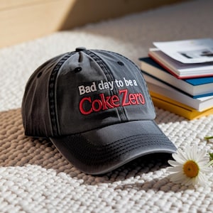 May include: A gray baseball cap with the text "Bad day to be a Coke Zero" embroidered in red and white. The cap is sitting on a white textured surface, with a stack of books in the background.