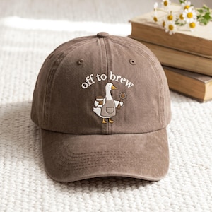 May include: A brown baseball cap featuring an embroidered graphic of a white goose wearing a grey apron, holding a coffee cup and a portafilter, with the white text "off to brew" arched above the goose in a simple, clean font style.