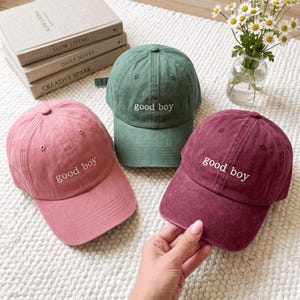 Può includere: Tre cappellini da baseball in rosa polvere, verde salvia e bordeaux scuro, ognuno con la scritta "good boy" ricamata in filo bianco sul pannello frontale. I cappellini hanno una corona strutturata, una visiera curva e un bottone in cima.