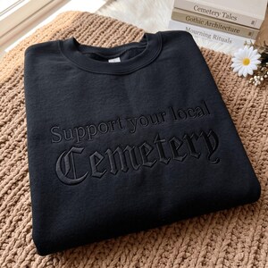 May include: A black crewneck sweatshirt featuring tonal black embroidery on the chest. The text reads Support your local Cemetery, with the word Cemetery displayed in a large, gothic-style font and the words Support your local in a smaller font.