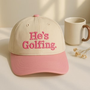May include: A beige baseball cap with a pink brim and a pink button on top. The cap features the embroidered text "He's Golfing" in pink. A beige mug with a brown handle is in the background.