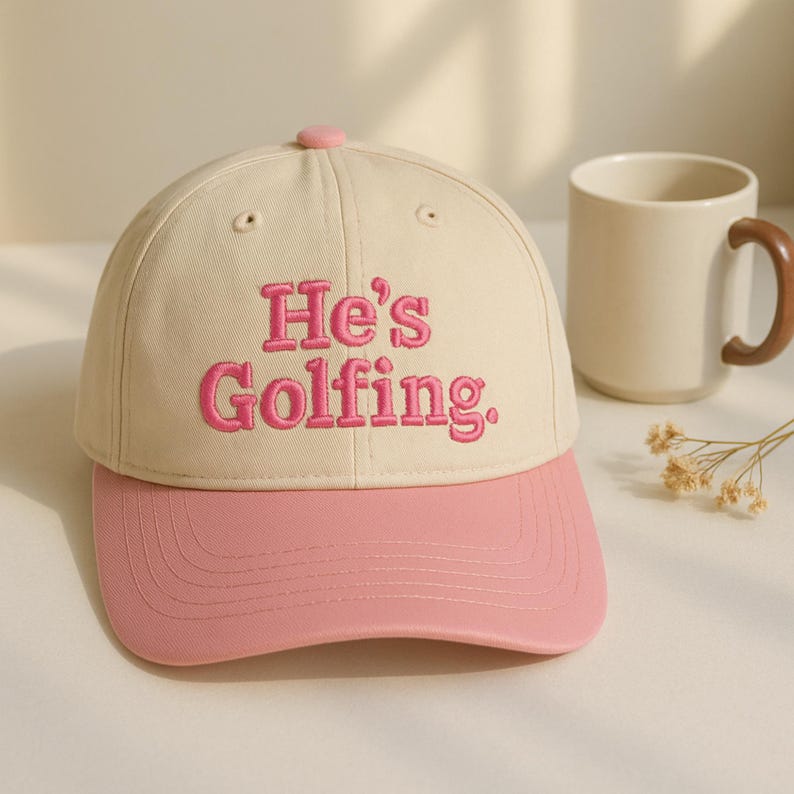 May include: A beige baseball cap with a pink brim and a pink button on top. The cap features the embroidered text "He's Golfing" in pink. A beige mug with a brown handle is in the background.