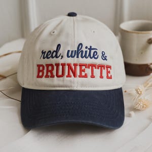May include: A baseball cap with a cream-colored crown and a navy blue brim. The cap features the embroidered text "red, white & BRUNETTE" in red and blue thread. A small navy blue button is on top.