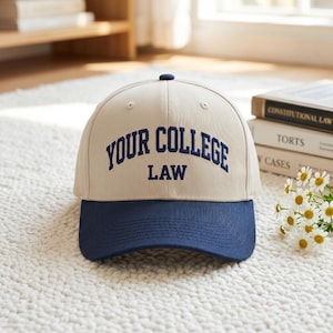 May include: A beige and navy blue baseball cap with the text "YOUR COLLEGE LAW" embroidered in navy blue. The cap has a curved brim and a button on top. The hat is sitting on a white, textured surface.