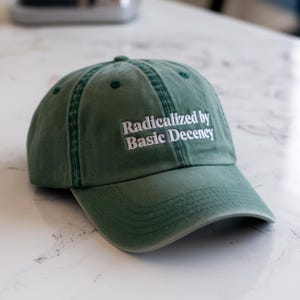 May include: A green baseball cap with the text "Radicalized by Basic Decency" embroidered in white.