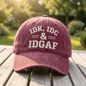 May include: A maroon baseball cap featuring white embroidered text that reads IDK, IDC & IDGAF. The cap has a structured crown, a curved brim with visible stitching, and a button on top, showcasing a washed fabric texture throughout the design.
