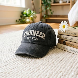 May include: A dark gray baseball cap with the word "ENGINEER" in white block letters and "EST. 2026" underneath. The cap sits on a white, textured rug next to a stack of books and a daisy.
