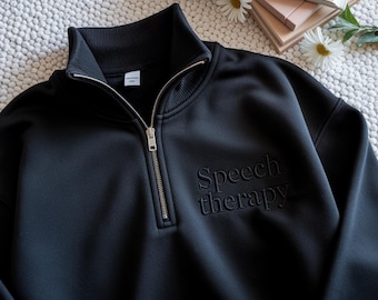 May include: Black quarter-zip sweatshirt with the embroidered text "Speech therapy". The sweatshirt has a high collar and a silver zipper. The garment is displayed on a textured white surface with a book and daisies in the background.
