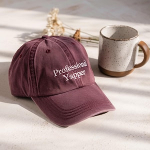 May include: A maroon baseball cap with the text "Professional Yapper" embroidered in white. A speckled white mug with a brown handle and base sits to the right of the cap. Dried flowers are in the background.
