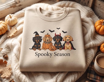 May include: A beige crewneck sweatshirt featuring a graphic of four dachshunds in Halloween costumes, including a witch, a pumpkin, a devil, and a vampire. Above the dogs are black bats, and below is the text Spooky Season in black font.