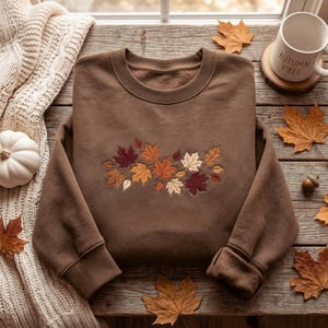 Puede incluir: Una sudadera marrón de cuello redondo con un bordado de hojas otoñales en tonos rojo, naranja, amarillo y crema en el centro del pecho. La prenda tiene mangas largas con puños acanalados y un cuello acanalado, confeccionada en tela suave.