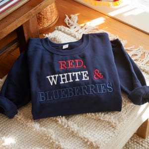 May include: A navy blue sweatshirt with the embroidered text "RED, WHITE & BLUEBERRIES" in red, white, and blue thread. The sweatshirt is folded on a cream-colored, textured surface.