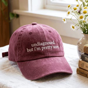 Può includere: Un cappellino da baseball color marrone scuro con struttura a sei pannelli e visiera curva. La parte anteriore presenta un testo ricamato in bianco che recita: "undiagnosed. but i'm pretty sure.". Il tessuto ha un aspetto leggermente sbiadito.